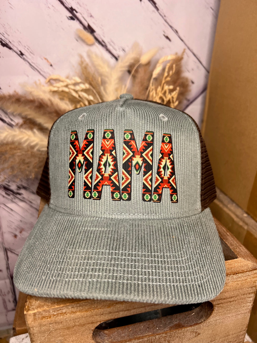 A corduroy trucker hat with 'MAMA' written in an Aztec pattern, featuring a mint green front and a brown mesh back.
