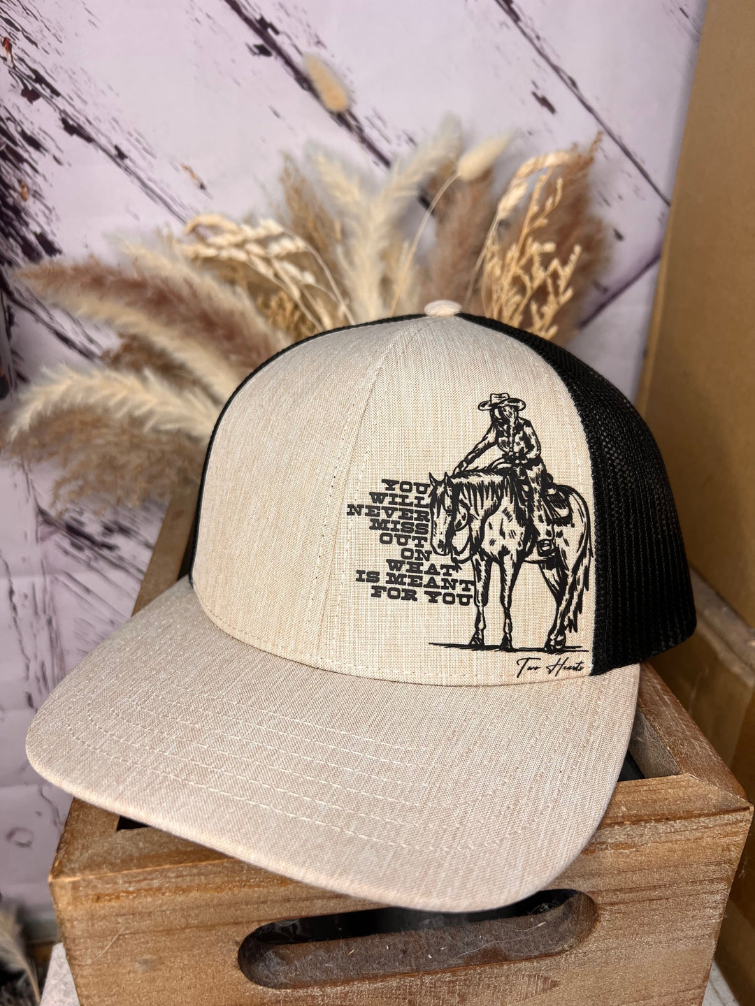 A beige and black ballcap with a graphic of a cowgirl on her horse and the text 'You will never miss out' printed on the side panel.