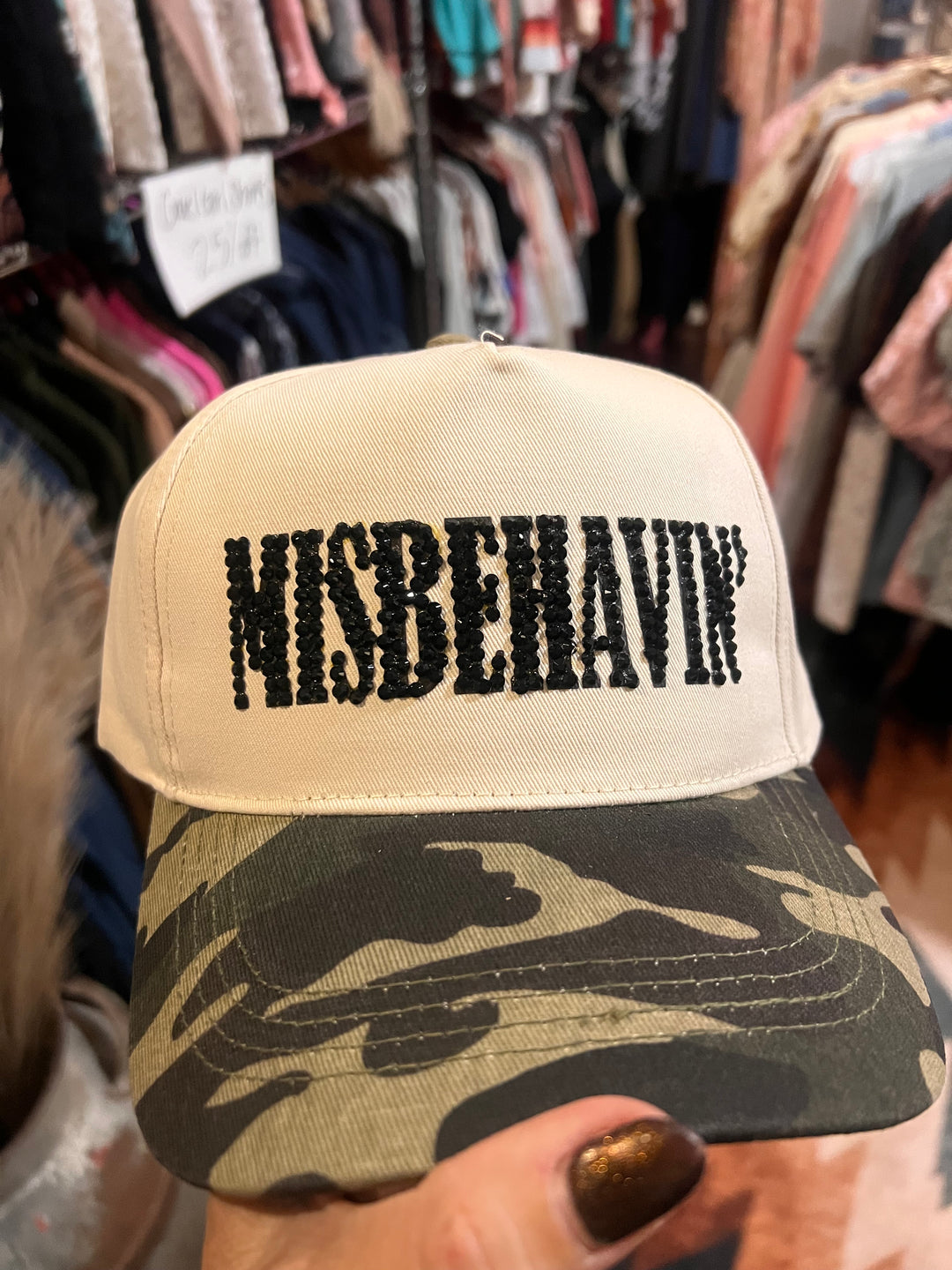A close-up of a baseball cap held by a hand, featuring a cream-colored front panel with the word 'MISBEHAVIN'' in black distressed lettering and a camouflage-patterned visor.