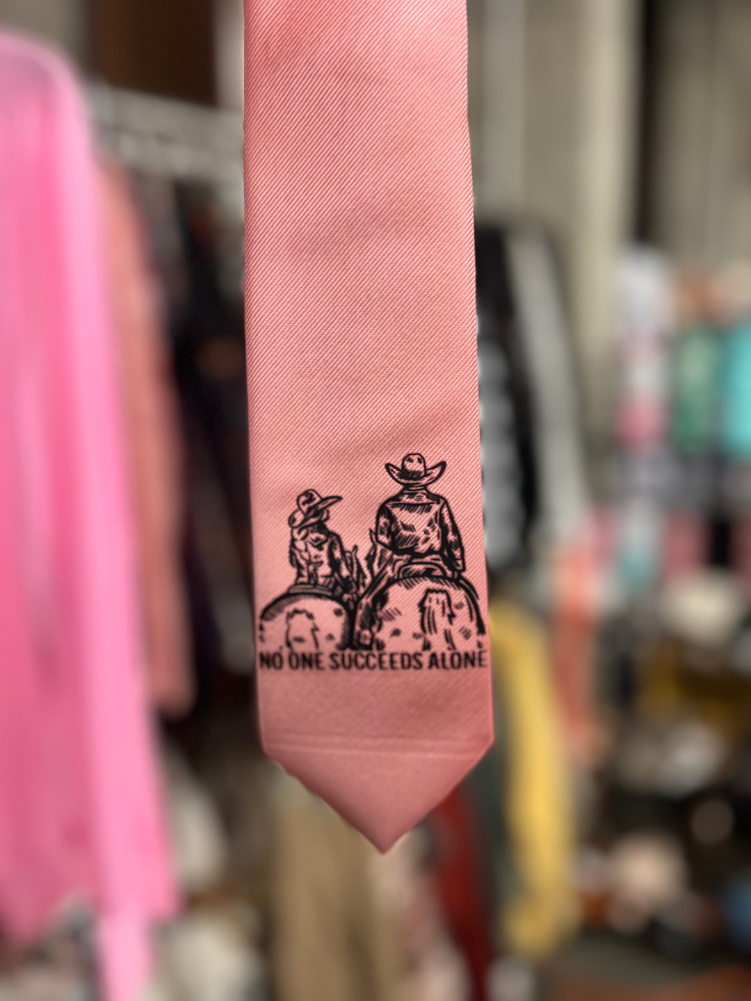 Pink tie with black graphic and text 'No One Succeeds Alone' in a store setting