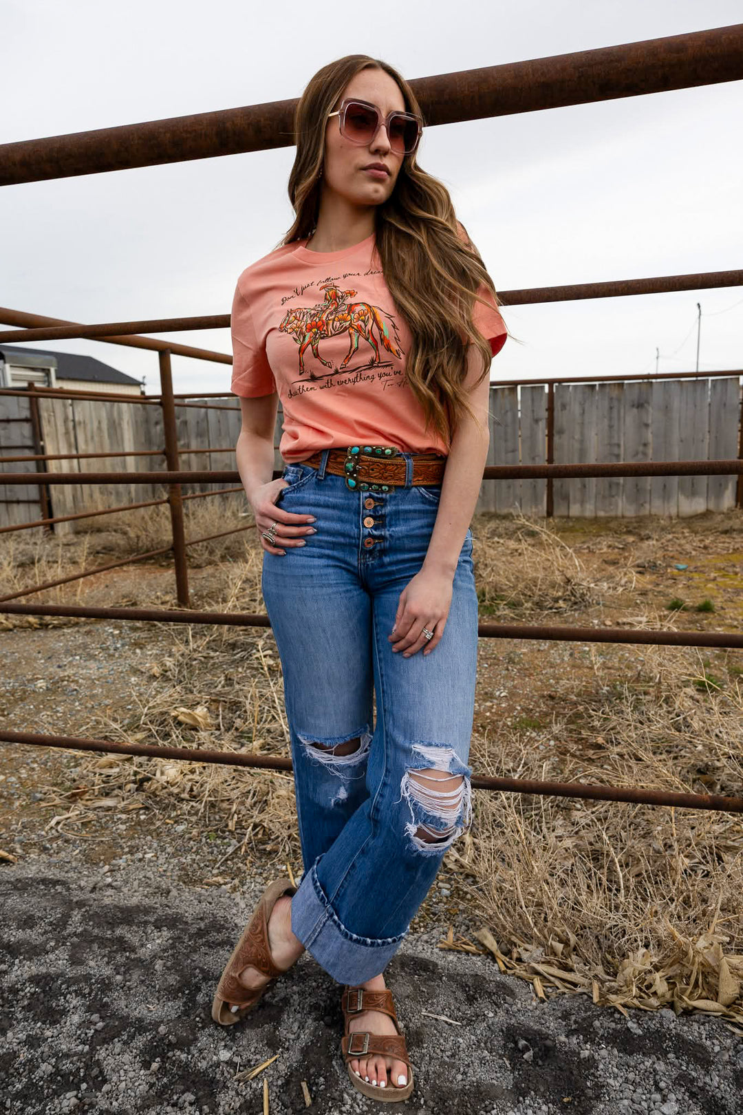 A woman standing outdoors wearing a pink graphic tee with a floral cowgirl design and the text 'CHASE YOUR DREAMS' on it, along with blue ripped jeans and brown sandals.