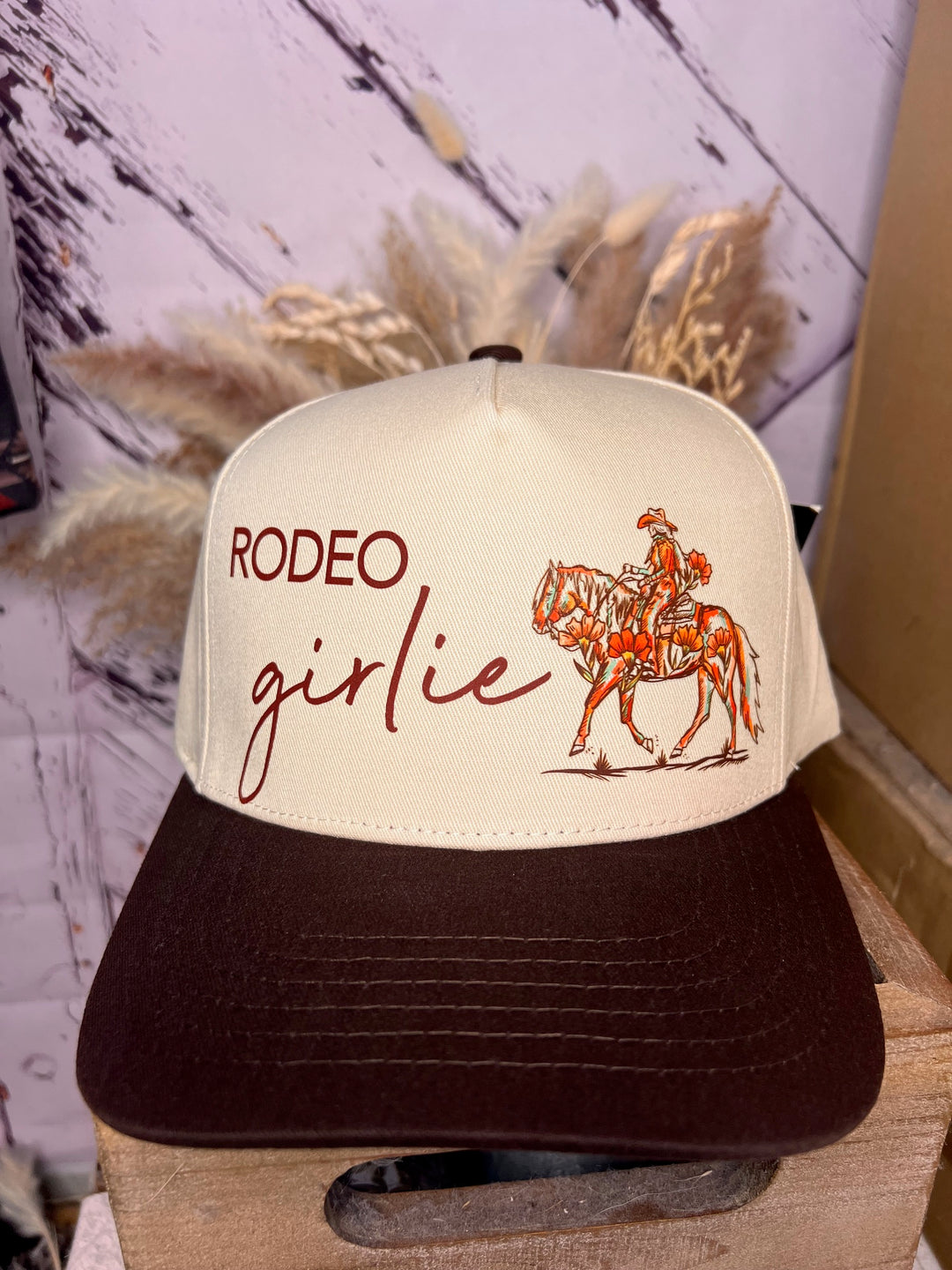 A beige and brown cowgirl cap with a graphic that includes the text 'RODEO girlie' and an illustration of a cowgirl riding a horse.