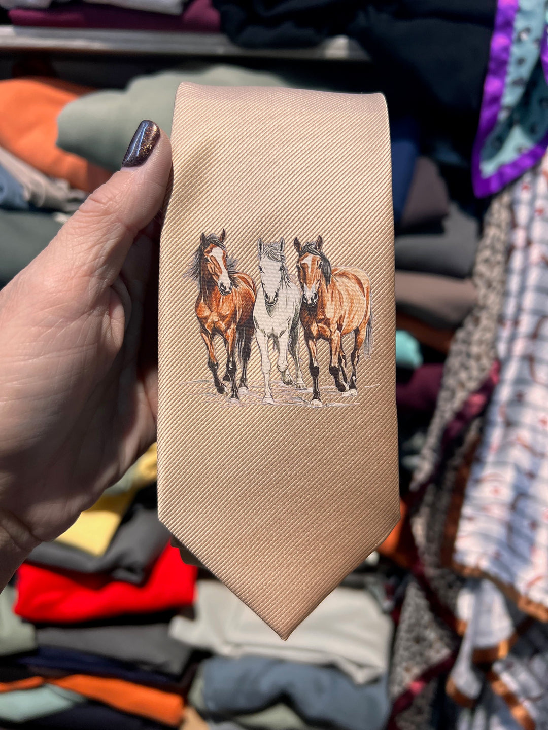 Beige belt with horse design held in front of a clothing rack.