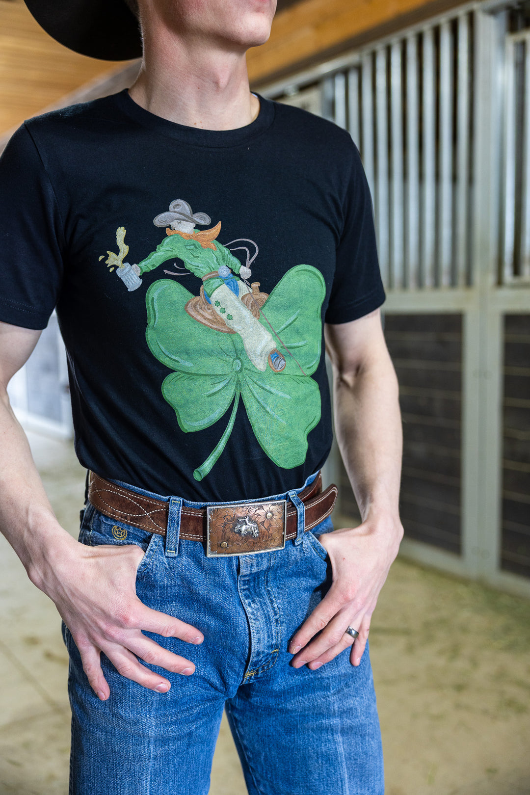 A person wearing a black graphic tee with a design of a cowboy bronc rider on a shamrock, paired with blue jeans and a brown belt.