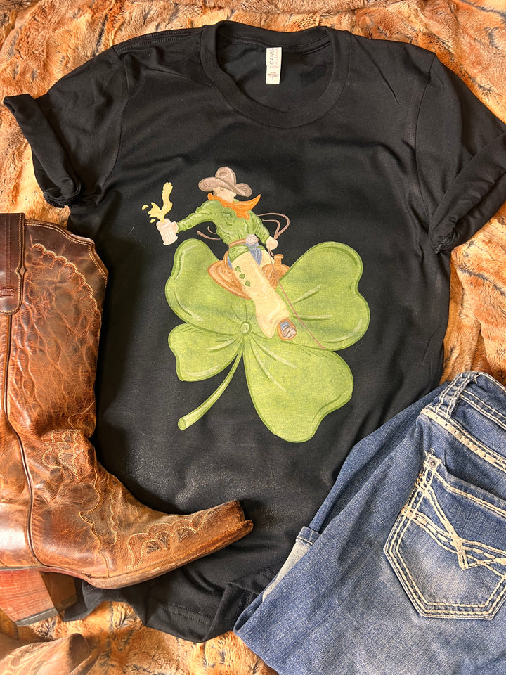 A black graphic tee with a design of a cowboy bronc rider on a shamrock, displayed alongside a pair of boots and jeans.