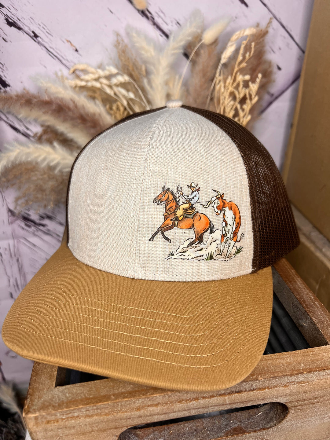 A beige and brown ballcap with a hand-drawn graphic of a cowboy on a horse ropes a steer, set against a wooden backdrop with a blurred floral pattern.