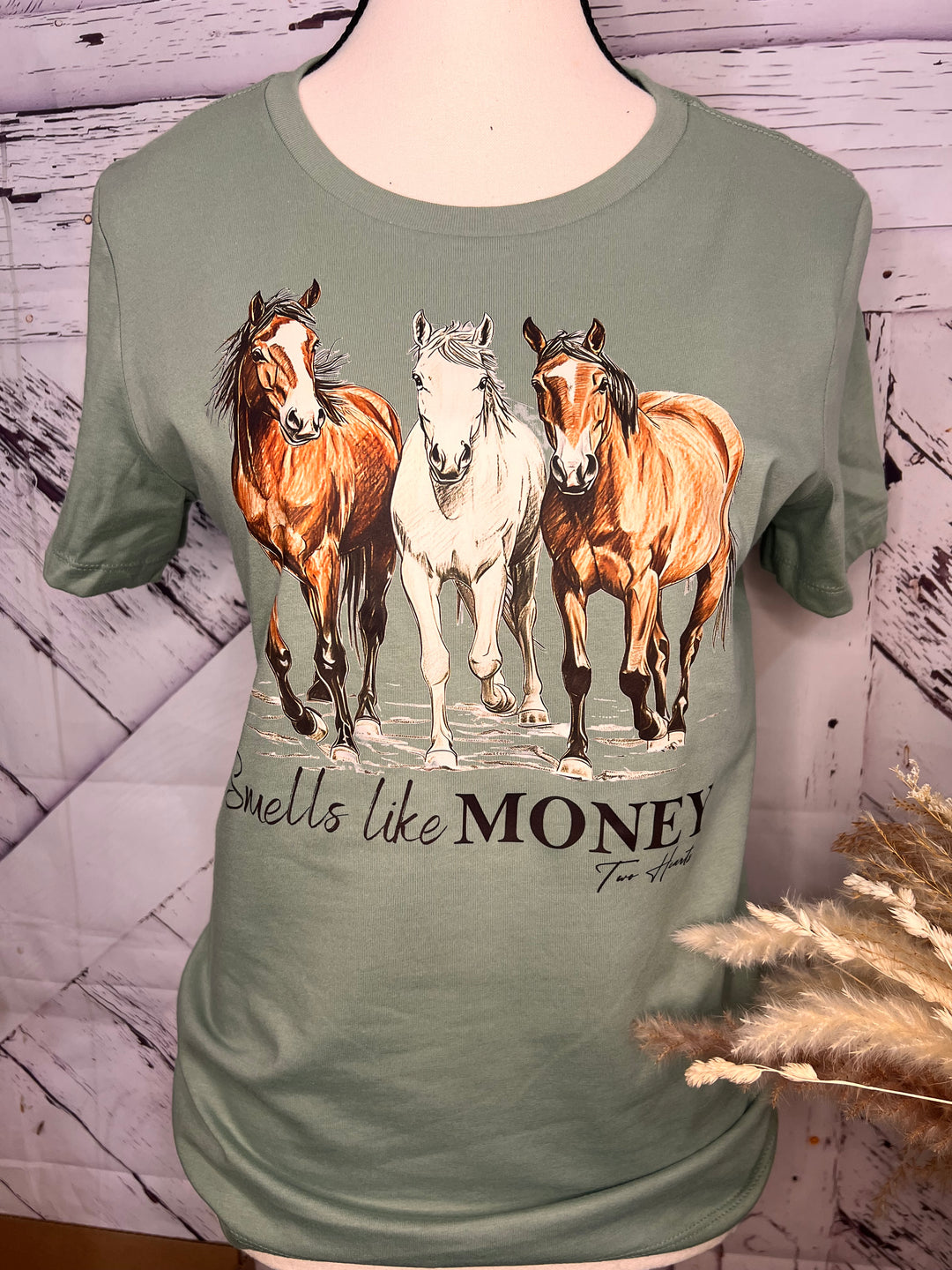 A sage green unisex cotton t-shirt with a graphic print of three horses trotting and the text 'Smells like MONEY'.