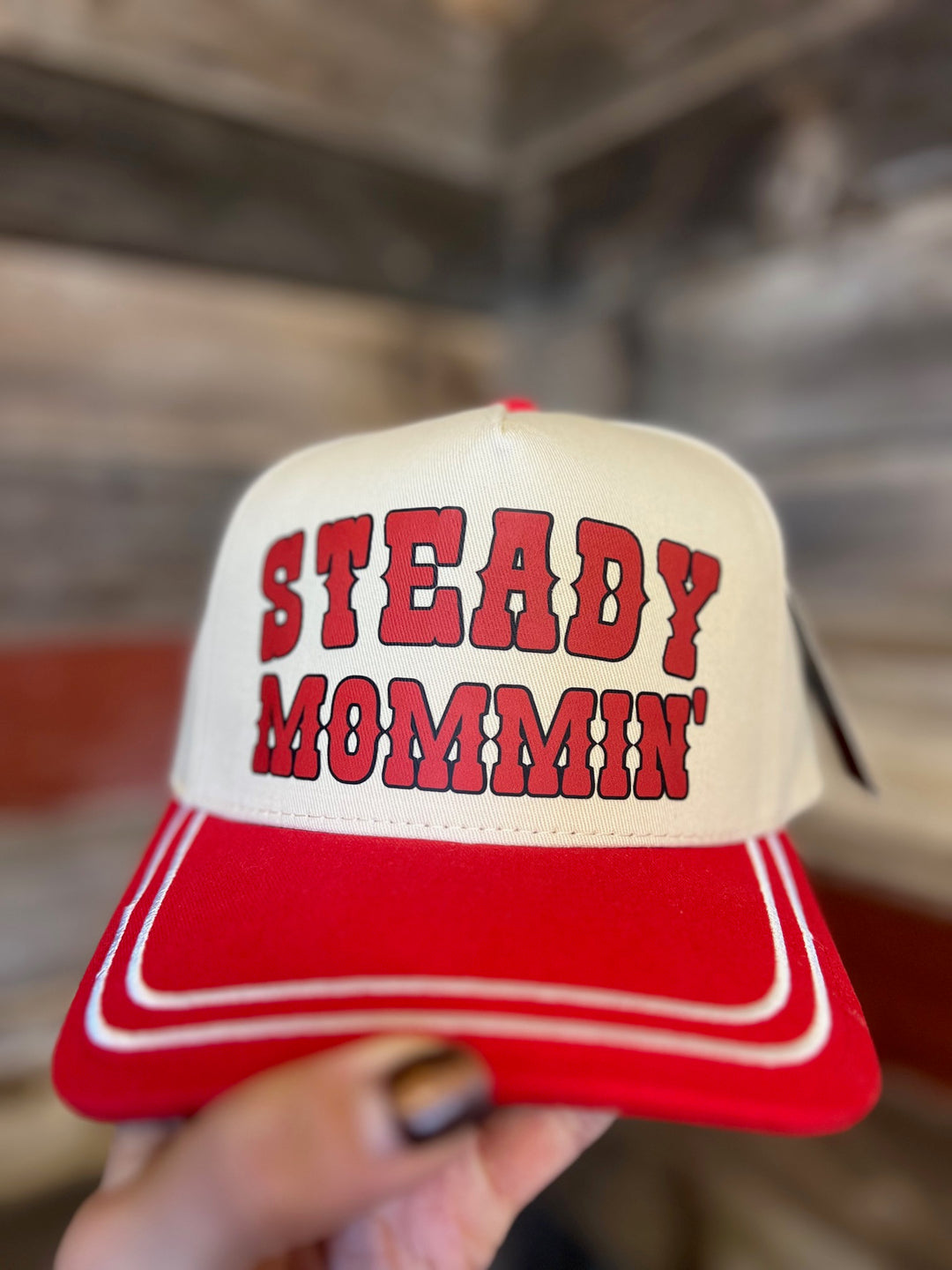 Red and white cap with 'STEADY MOMMIN'' text held by a hand against a blurred background
