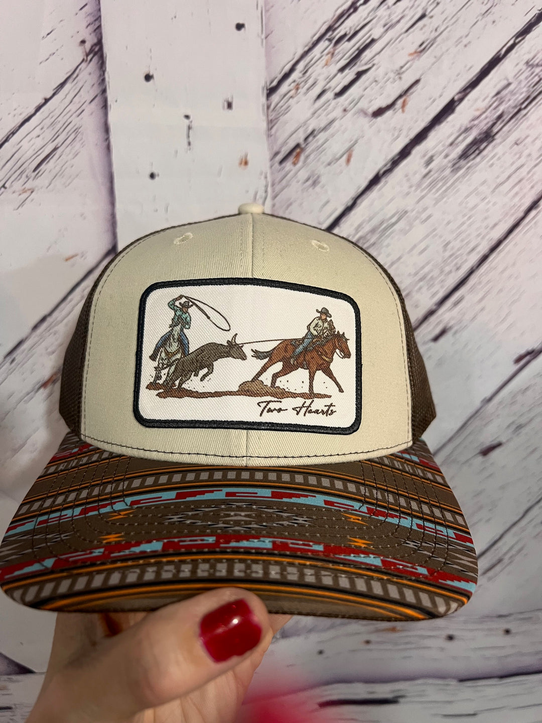 A beige and multicolored ballcap with a hand-drawn graphic of two people team roping on a stitch patch on the front.