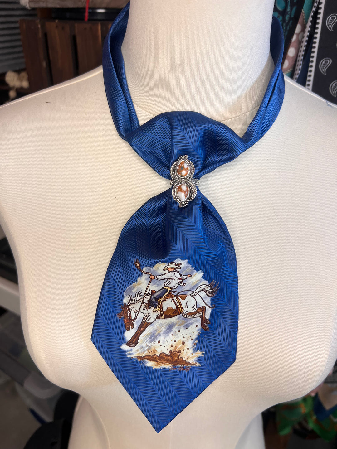 A royal blue necktie styled as a collar, featuring a printed equestrian scene on the front and two silver-toned decorative studs with pinkish crystal centers at the knot, displayed on a white mannequin torso.