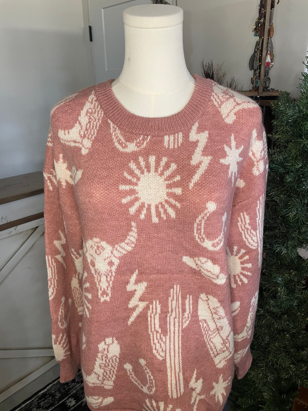 A dusty pink knit sweater with a cream western-themed pattern featuring cacti, a bull skull, boots, and lightning bolts, displayed on a white mannequin torso.