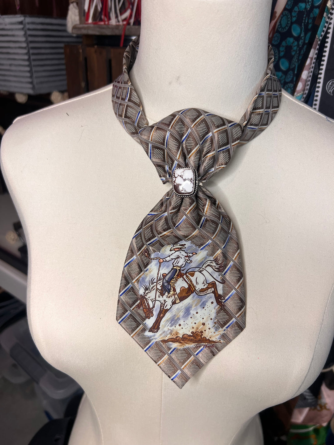 A patterned necktie displayed on a white mannequin torso. The tie features a gray and white basketweave texture with a central rhinestone knot and a western-themed printed section on the front blade showing a cowboy on horseback.