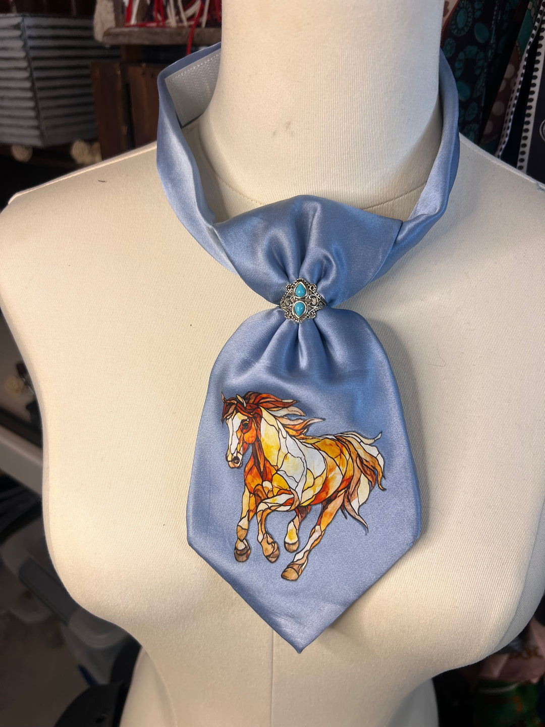 A light blue satin necktie styled on a cream mannequin torso, featuring a detailed illustration of a galloping horse in orange, white, and brown on the front, and a small decorative brooch at the knot.