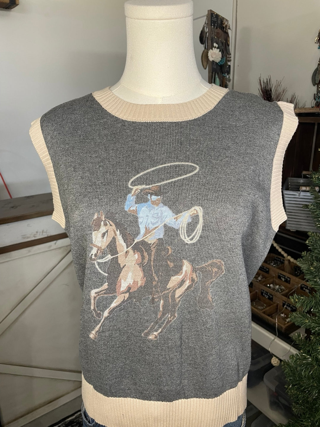 A gray sleeveless knit sweater vest displayed on a mannequin torso, featuring a cowboy riding a horse and throwing a lasso graphic in beige, blue, and brown tones, with cream ribbed trim.