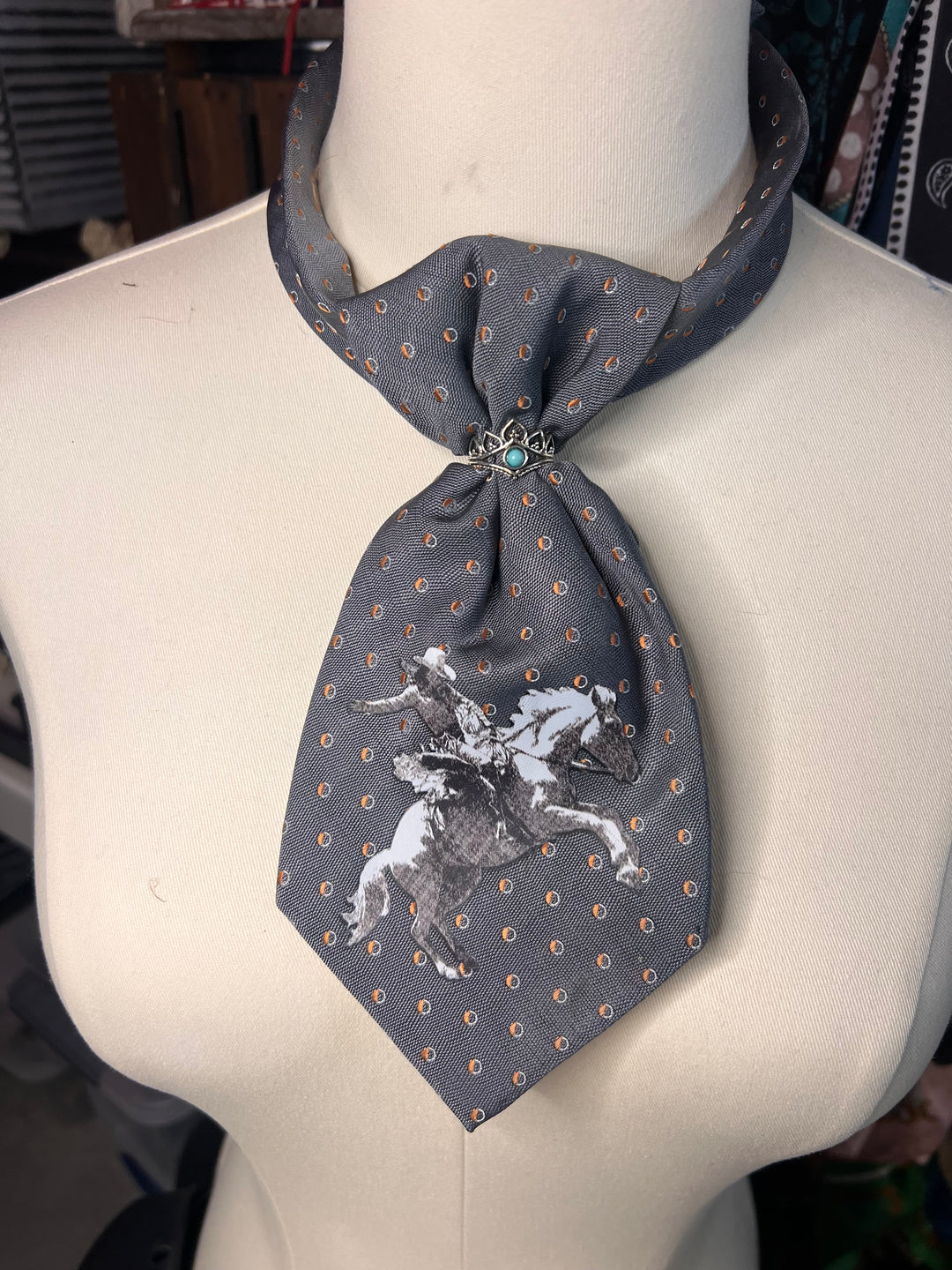 A gray silk necktie with a white horse graphic and gold dotted pattern, displayed on a cream-colored mannequin torso.