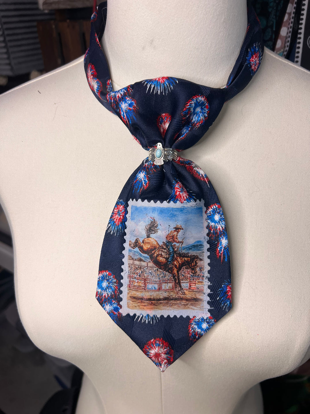 A dark navy necktie styled on a cream mannequin torso. The tie features a red, white, and blue fireworks pattern on the edges and a central rectangular stamp-like graphic depicting a cowboy riding a bucking bull.