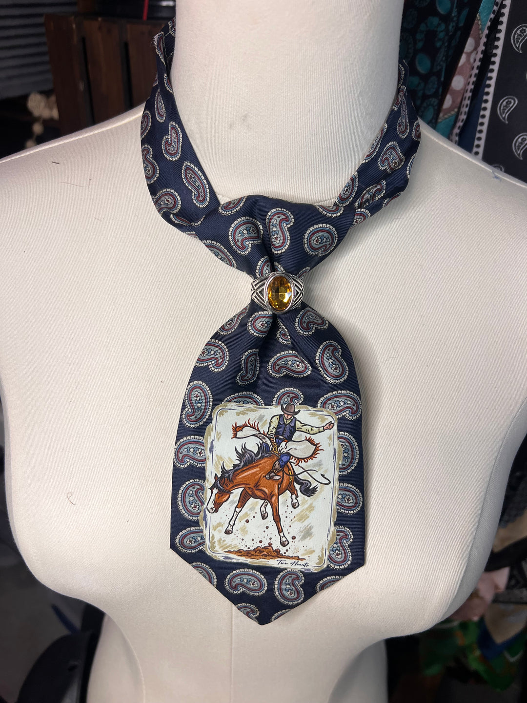 A novelty necktie displayed on a white mannequin torso. The tie features a navy blue paisley pattern with a central printed illustration of a cowboy riding a bucking brown horse on a white background, finished with a decorative gold-toned metal ring at the knot.