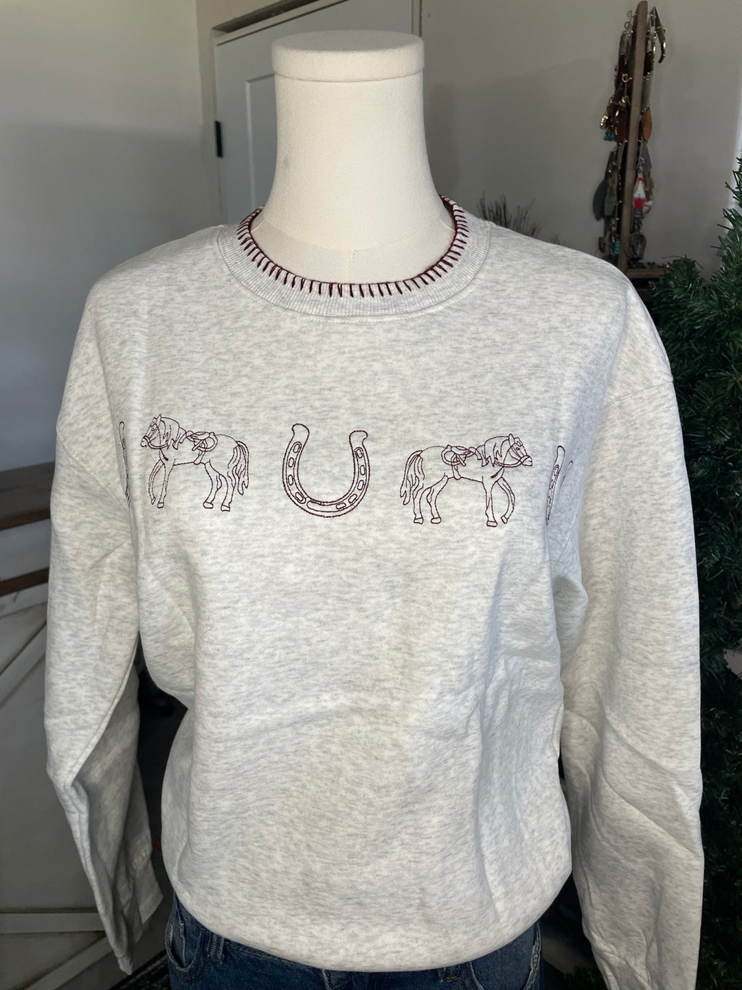 A light heather gray crewneck sweatshirt displayed on a white mannequin torso. The sweatshirt features embroidered brown designs across the chest, including two horses and a horseshoe.