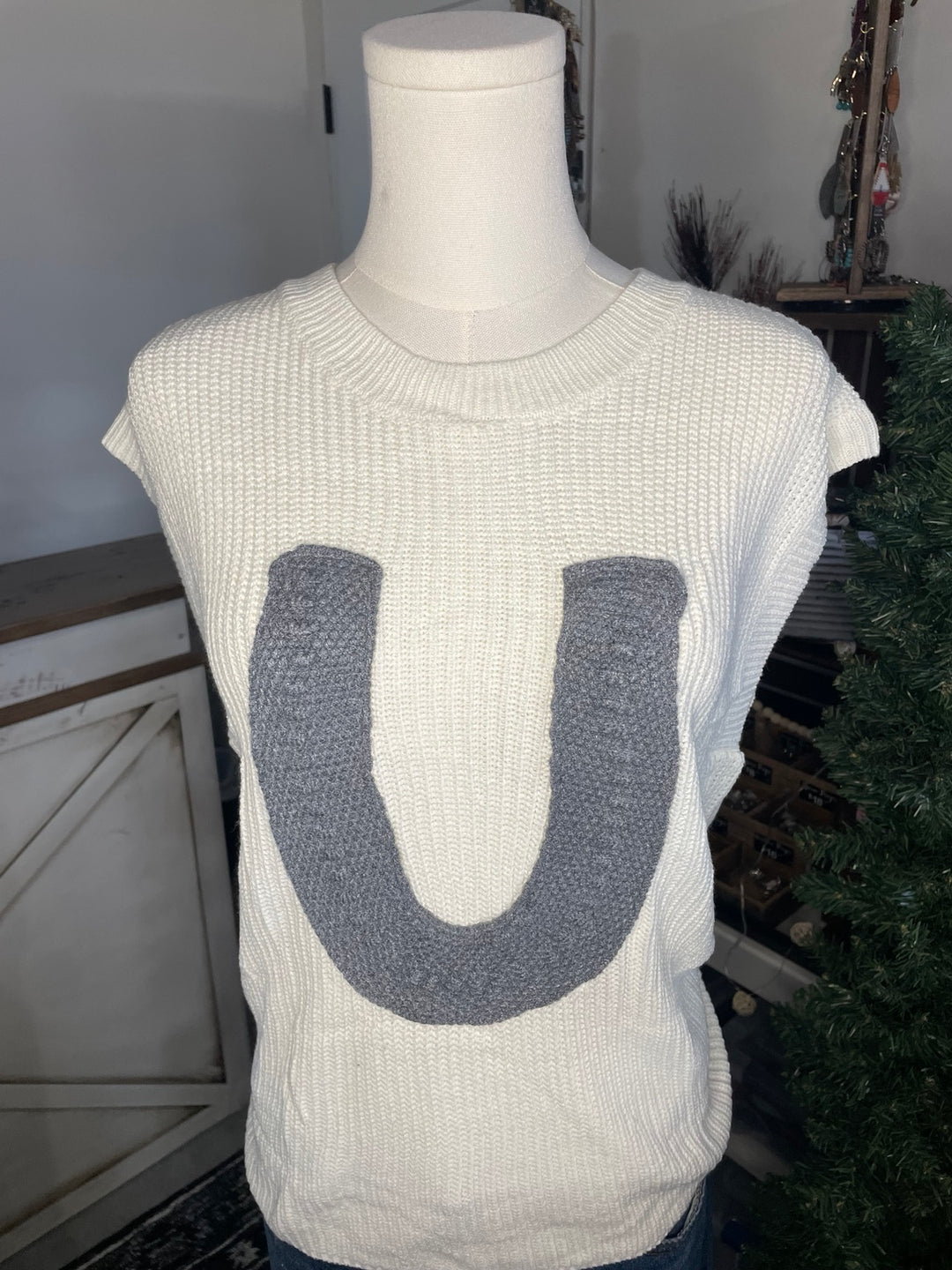 An off-white knit sweater vest displayed on a white mannequin torso, featuring a large gray knitted letter 'U' on the front.