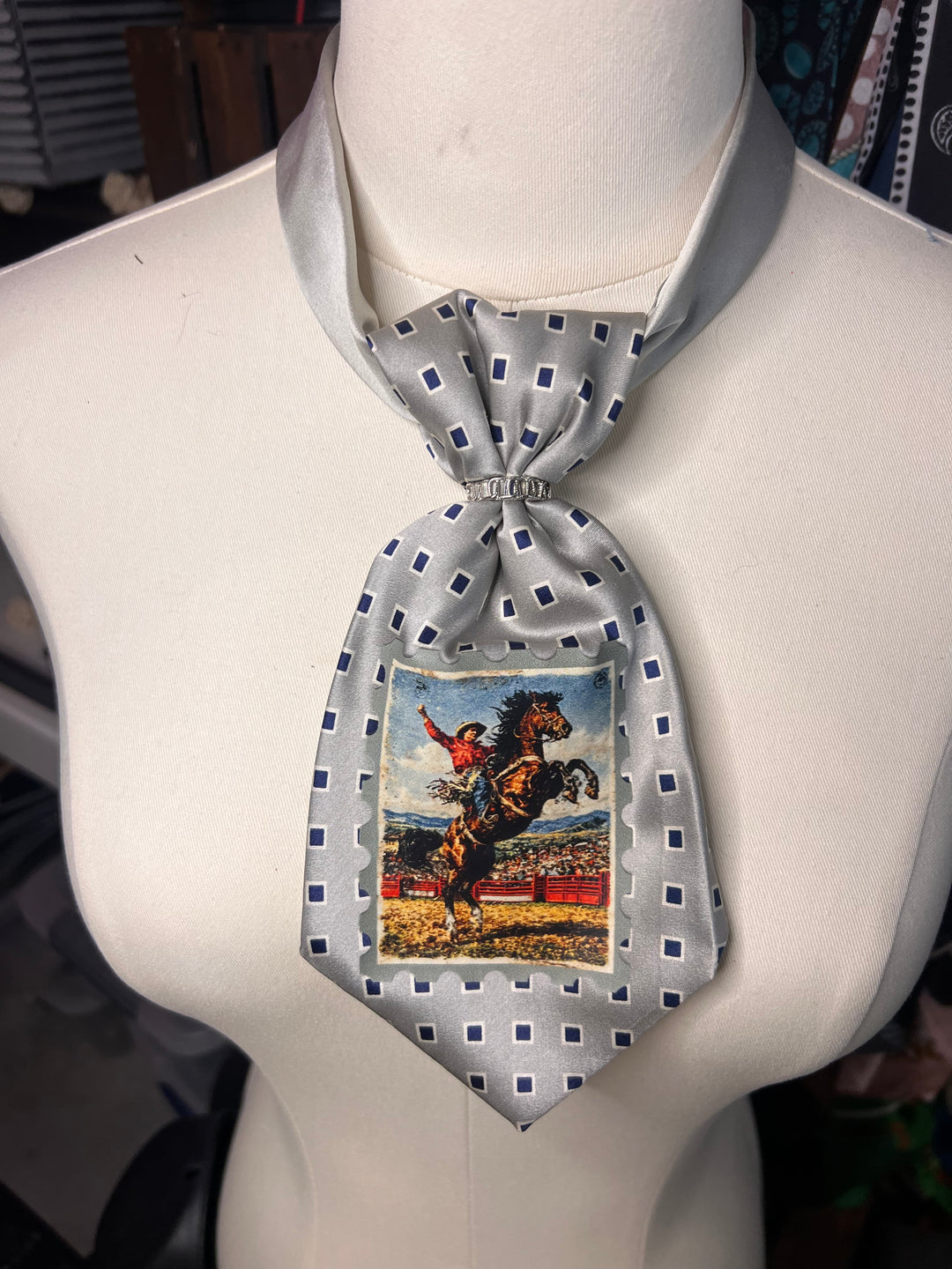 A light gray silk-like necktie with a blue geometric pattern and a central printed image of a rearing horse, displayed on a white mannequin torso.
