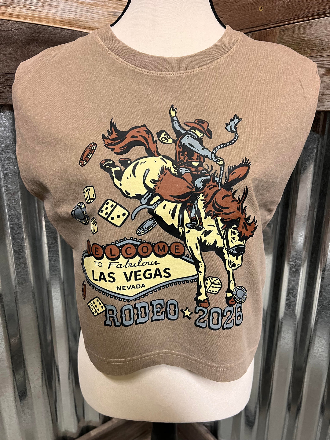 A cropped tan t-shirt displayed on a mannequin torso, featuring a large graphic print of a cowboy riding a bucking horse with dice and the text &