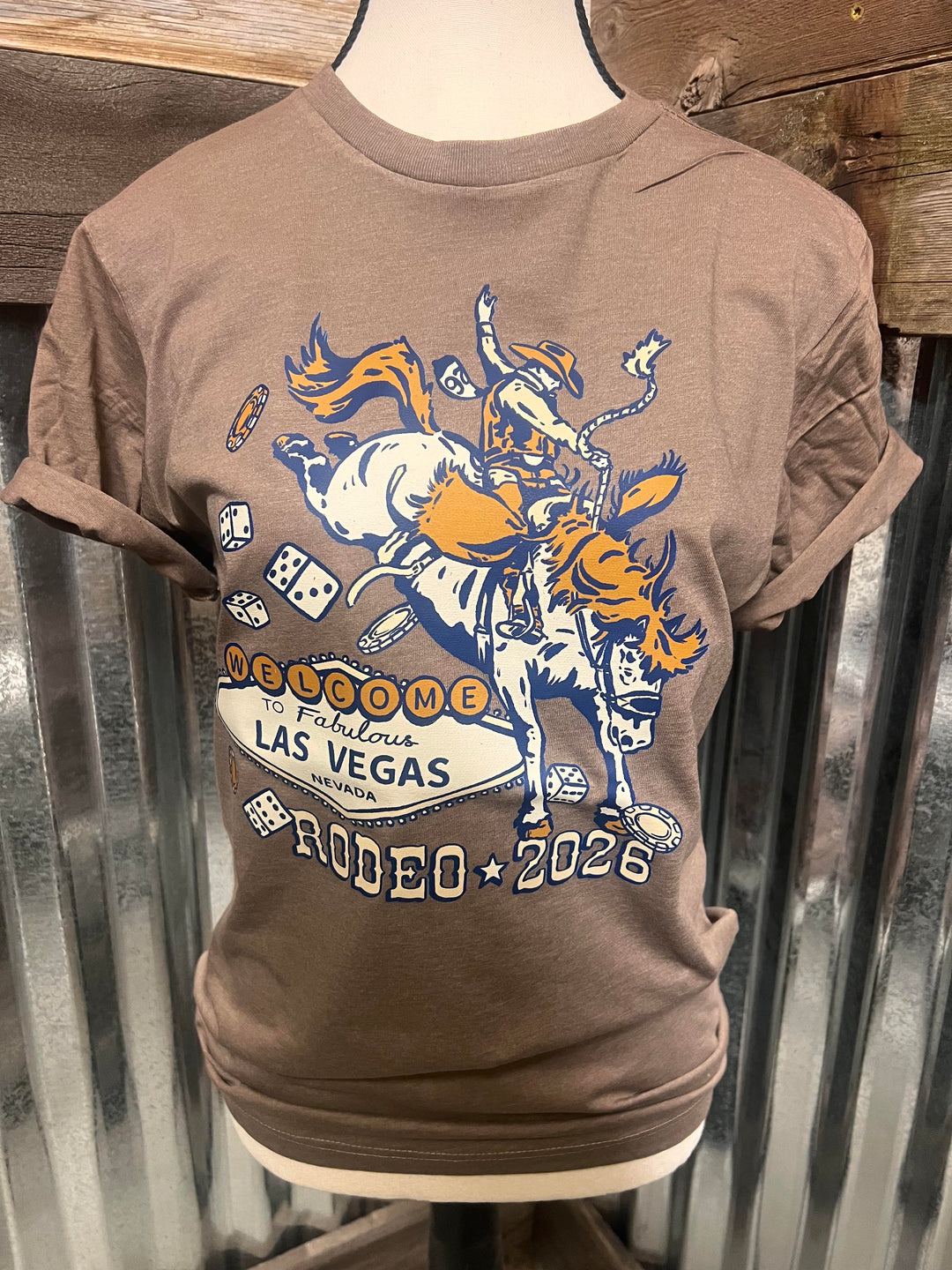 A brown short-sleeve graphic T-shirt displayed on a mannequin torso against a corrugated metal and wood backdrop. The shirt features a large front graphic with a cowboy riding a bucking bull, dice, and the text 'Welcome to Fabulous Las Vegas 2026' and 'Rodeo'.