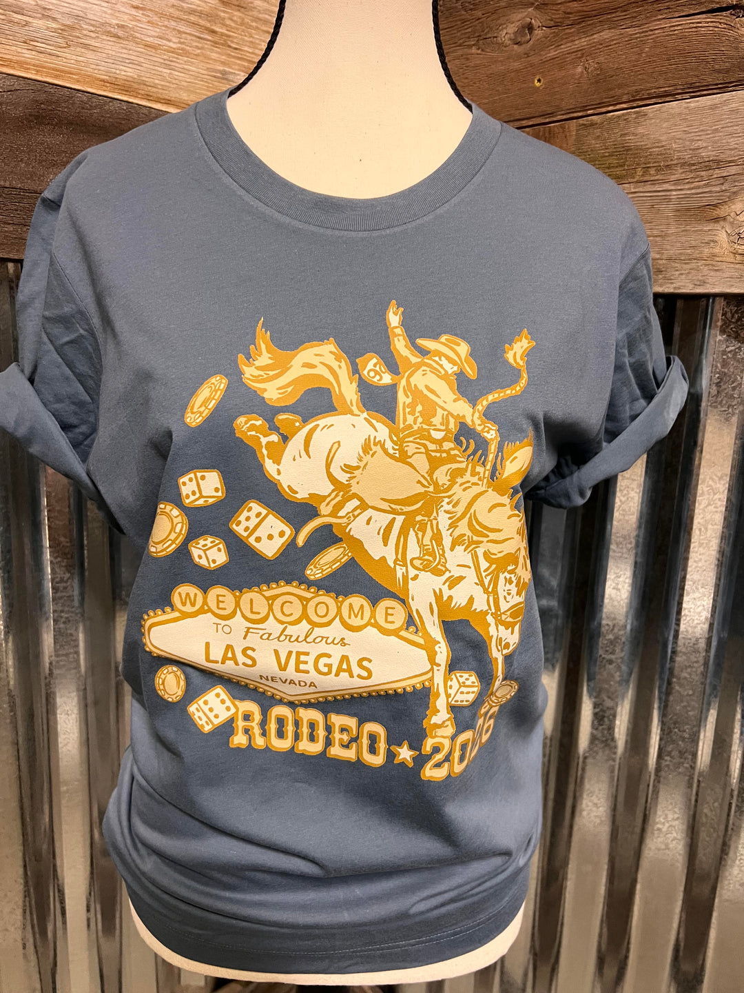 A slate blue t-shirt displayed on a mannequin torso, featuring a large yellow graphic print of a cowboy riding a bucking bronco with 'Welcome to Fabulous Las Vegas Rodeo 2026' and playing card designs.