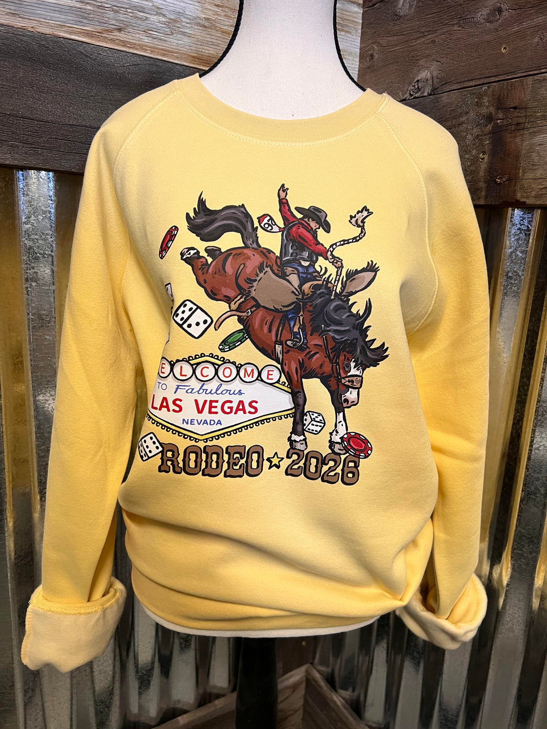 A pale yellow long-sleeve sweatshirt displayed on a mannequin torso, featuring a large western-themed graphic on the front with a cowboy on a bucking horse, dice, and the text &