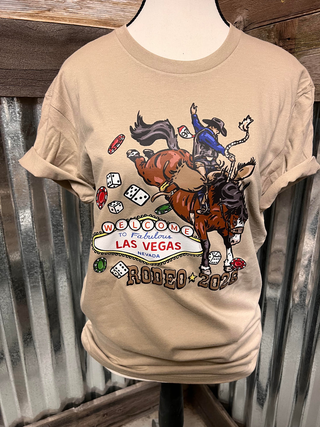 A beige graphic t-shirt displayed on a mannequin torso against a corrugated metal and wood background. The graphic features a cowboy riding a bucking bronco with dice, poker chips, and a 'Welcome to Fabulous Las Vegas Rodeo 2026' sign.