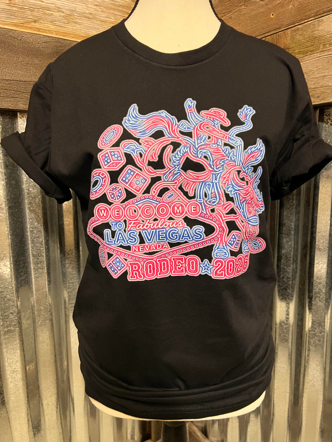 A black short-sleeve graphic T-shirt displayed on a mannequin torso against a corrugated metal and wood background. The shirt features a large front graphic in red and blue outline art with the text 'WELCOME to LAS VEGAS RODEO 2025'.