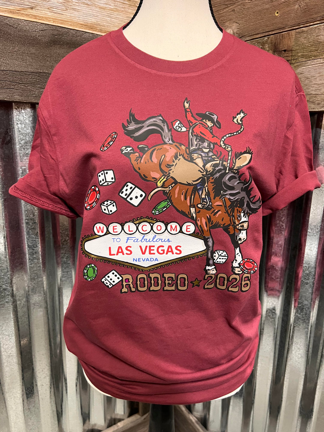 A maroon short-sleeve graphic t-shirt displayed on a mannequin torso against a rustic corrugated metal and wood background. The graphic features a cowboy riding a horse with rodeo and casino elements, and the text 'Welcome to Fabulous Las Vegas Nevada Rodeo 2026'.
