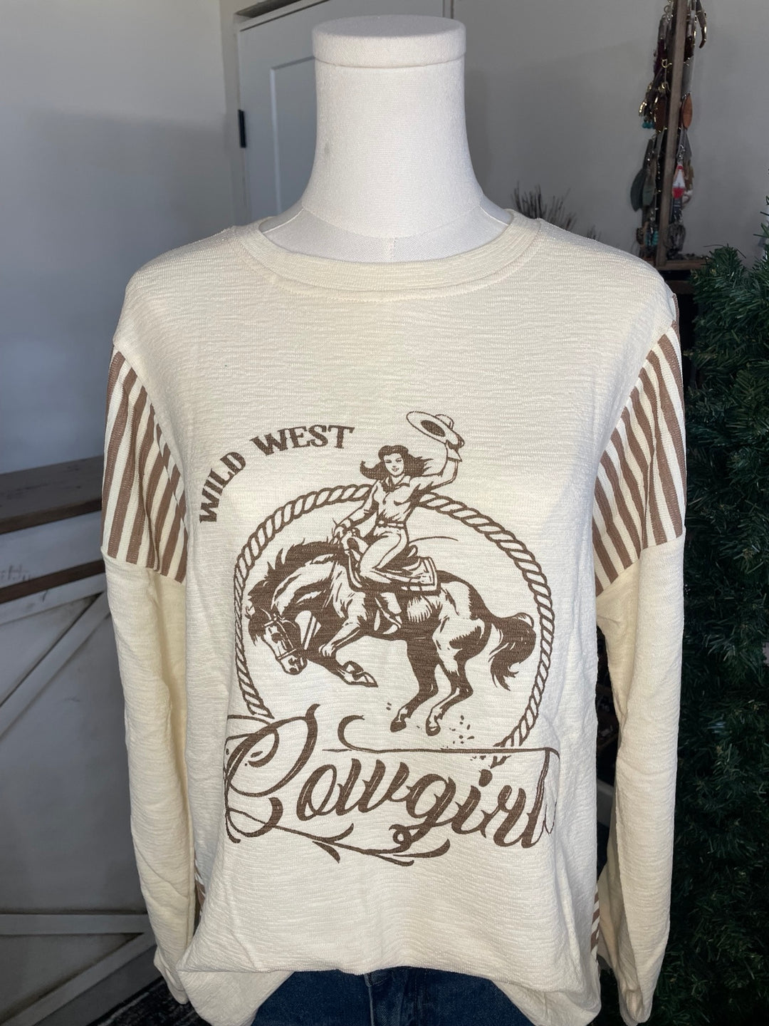 Cream long-sleeve graphic shirt on a mannequin torso with brown western print reading 'Wild West Cowgirls' and a cowboy riding a horse, plus brown striped sleeve accents.