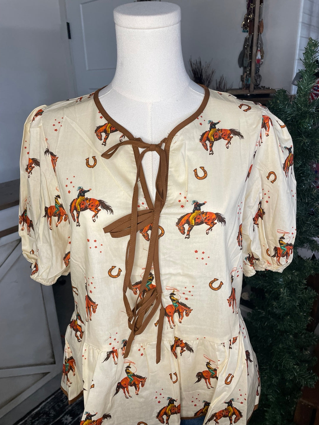 A cream-colored short-sleeve blouse with a brown tie-neck detail and an all-over print of brown horses, riders, horseshoes, and red splatters, displayed on a white mannequin torso.