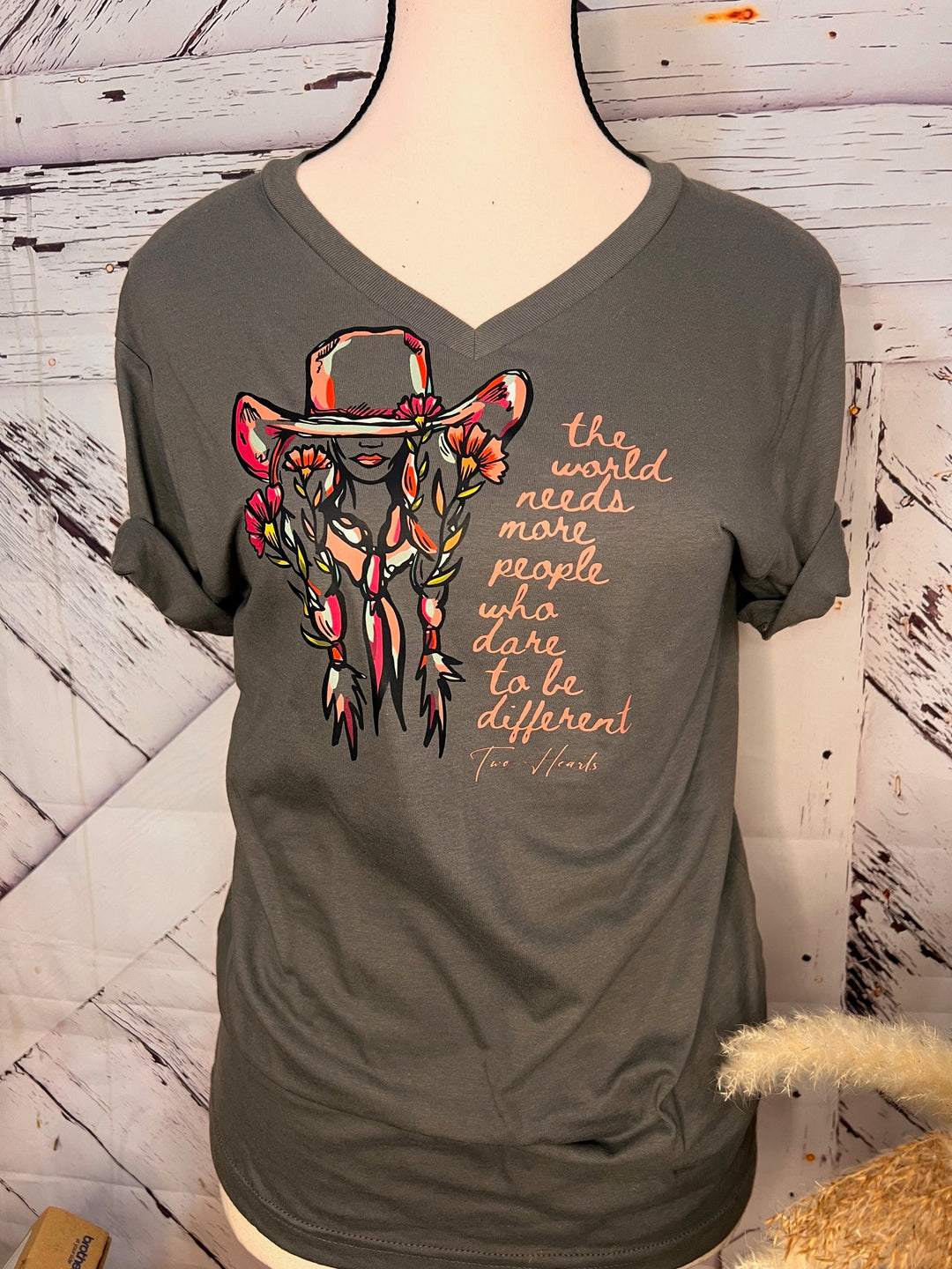 A person wearing a pink floral cowgirl tee with a quote graphic, paired with blue ripped jeans and a black jacket, sitting on a gate with a landscape in the background.