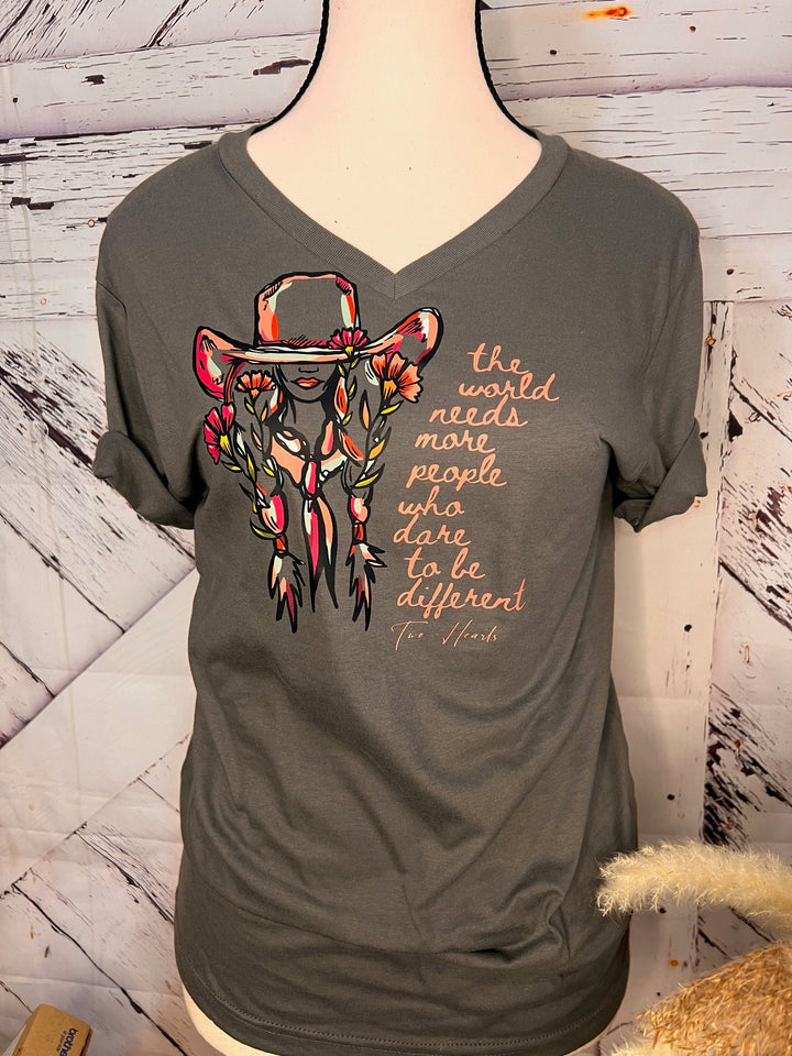 A person wearing a pink floral cowgirl tee with a quote graphic, paired with blue ripped jeans and a black jacket, sitting on a gate with a landscape in the background.