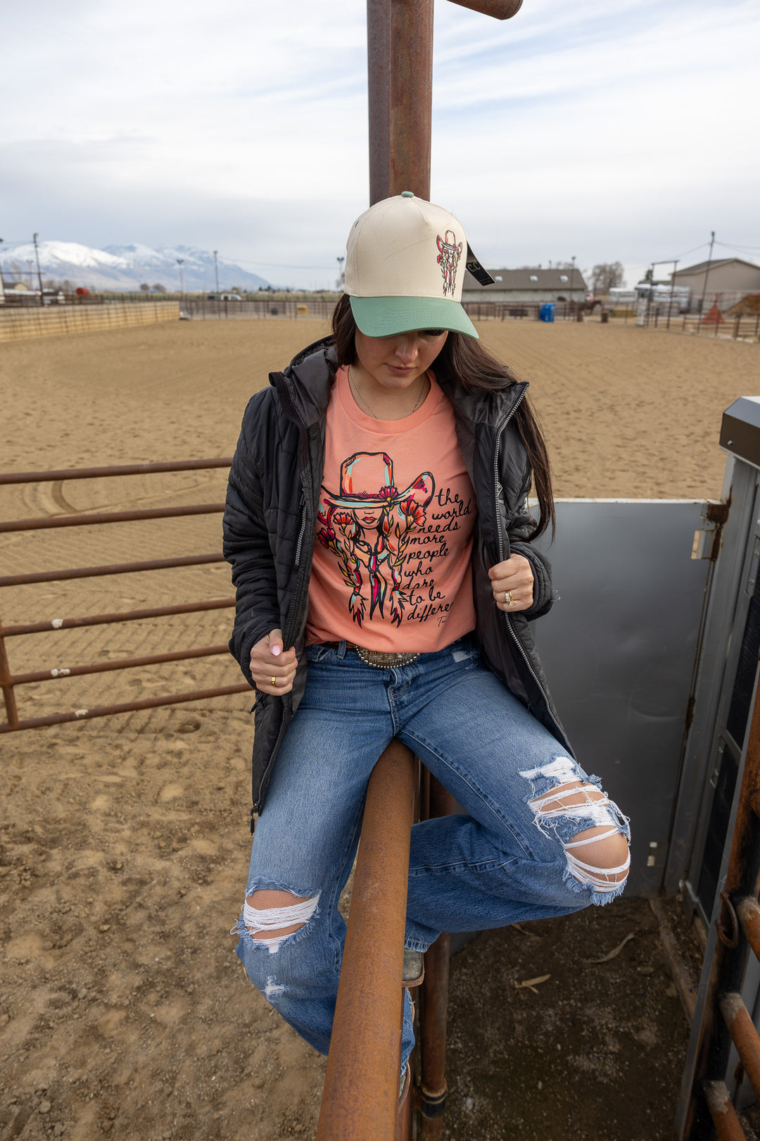 A person wearing a pink floral cowgirl tee with a quote graphic, paired with blue ripped jeans and a black jacket, sitting on a gate with a landscape in the background.