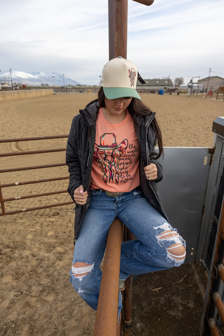 A person wearing a pink floral cowgirl tee with a quote graphic, paired with blue ripped jeans and a black jacket, sitting on a gate with a landscape in the background.