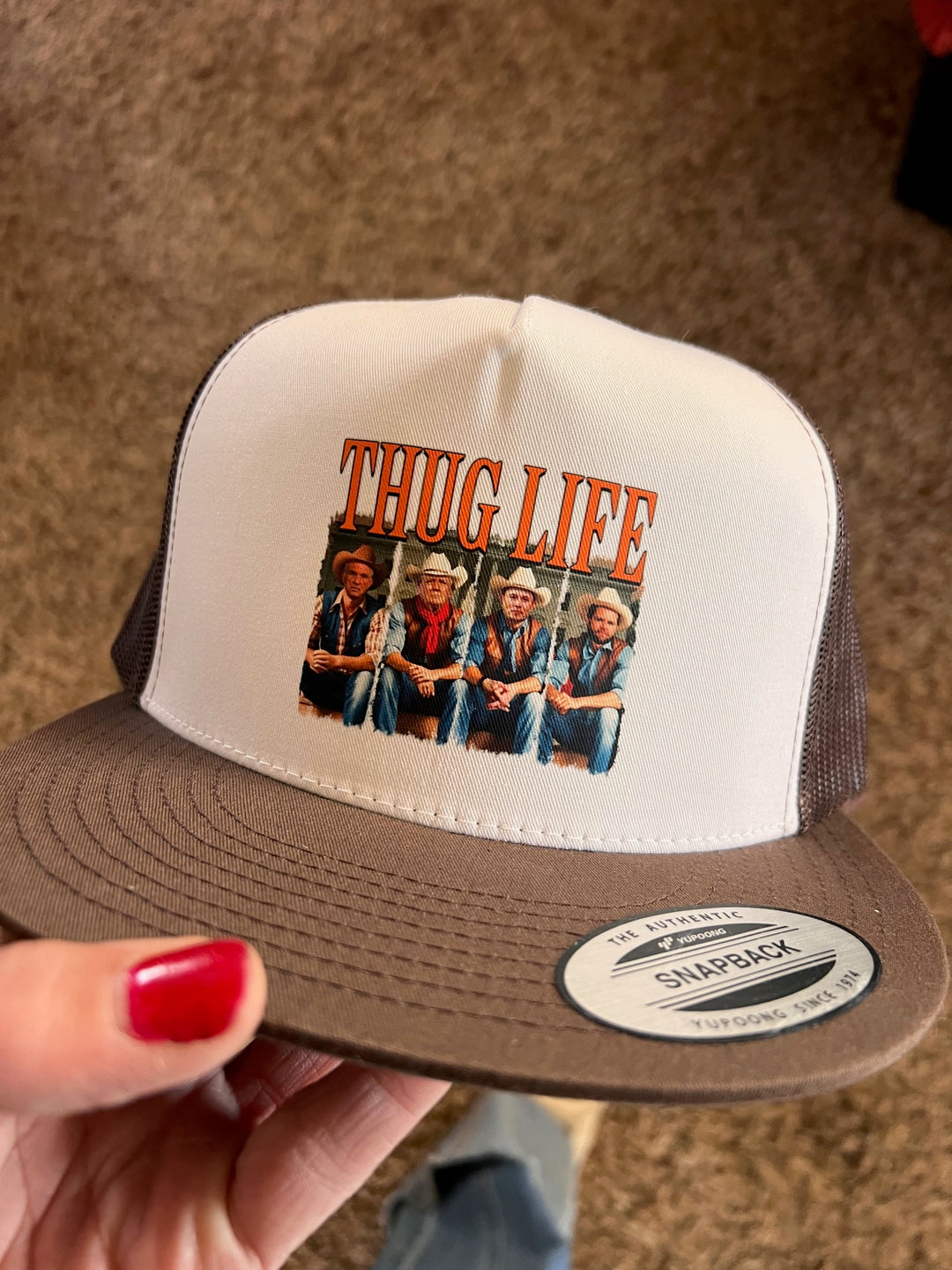 A beige and white ballcap with a graphic print of a phrase 'THUG LIFE' and images of individuals underneath it. The cap is held by a person with red nail polish.