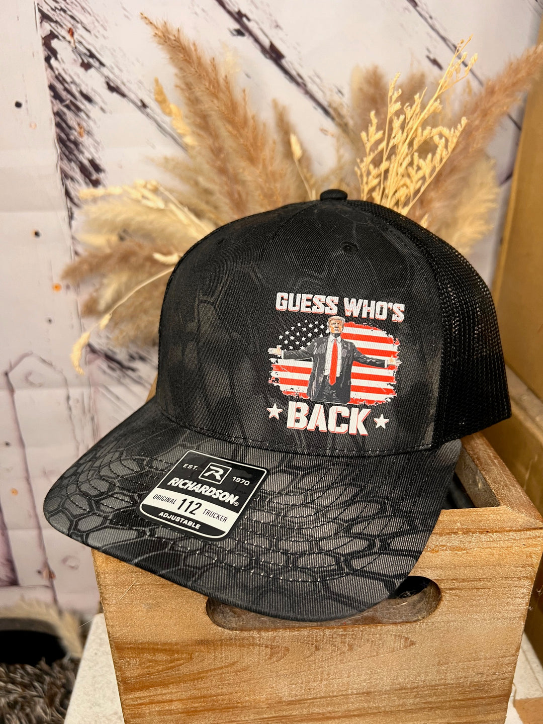 A black camo pattern ballcap with a design featuring Donald Trump and the text 'Guess who's back' on the front.