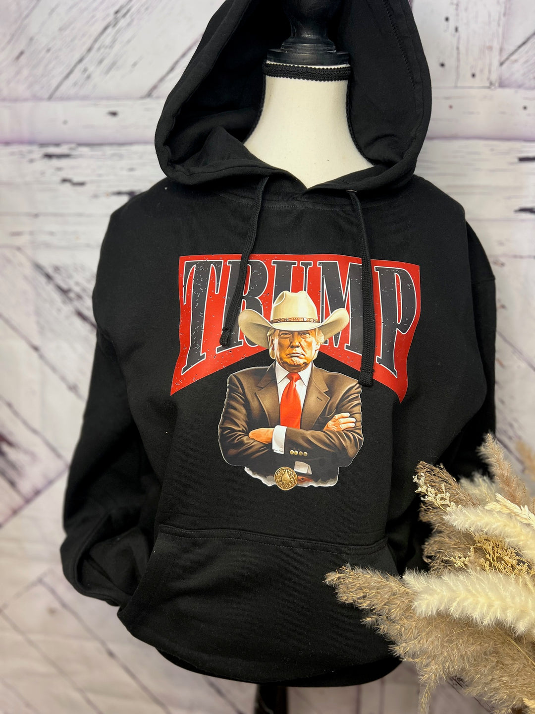 A black hoodie with a graphic print of a cartoon Donald Trump wearing a cowboy hat, with the text 'TRUMP' above the image.