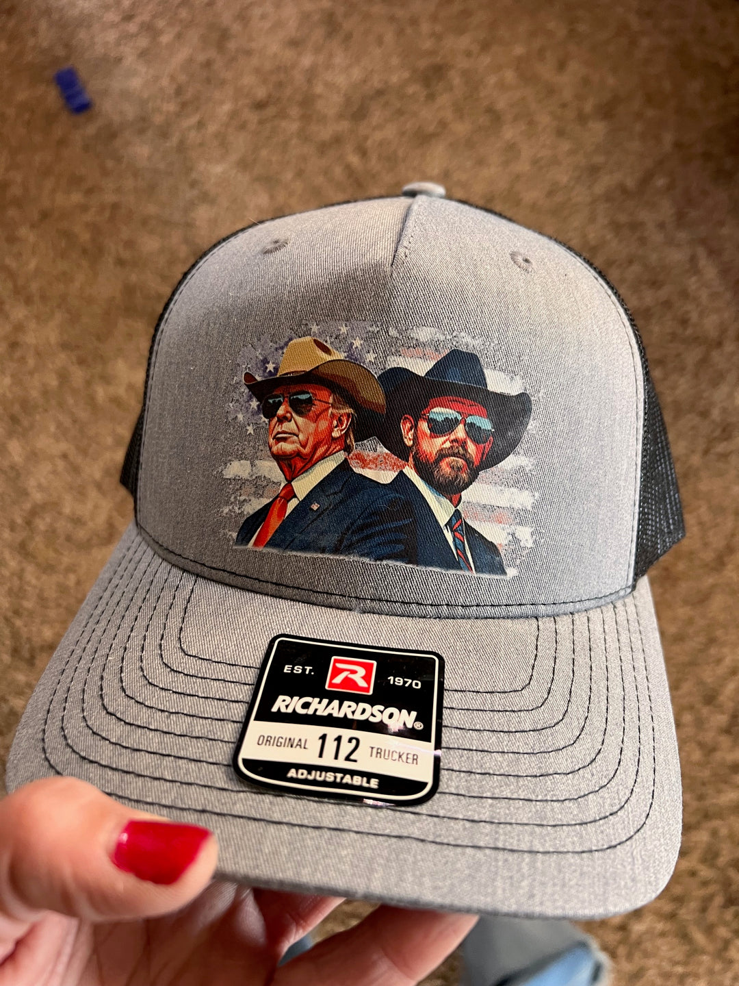 A gray and black ballcap with an illustration of Donald Trump and JD Vance wearing cowboy hats, set against a backdrop of the American flag.