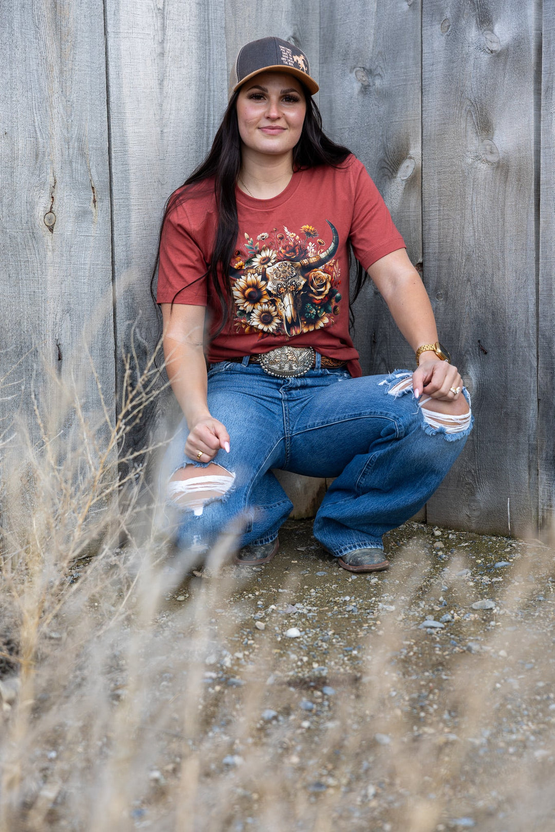 Woman sitting against a wooden wall wearing a red t-shirt with a graphic design and blue jeans.