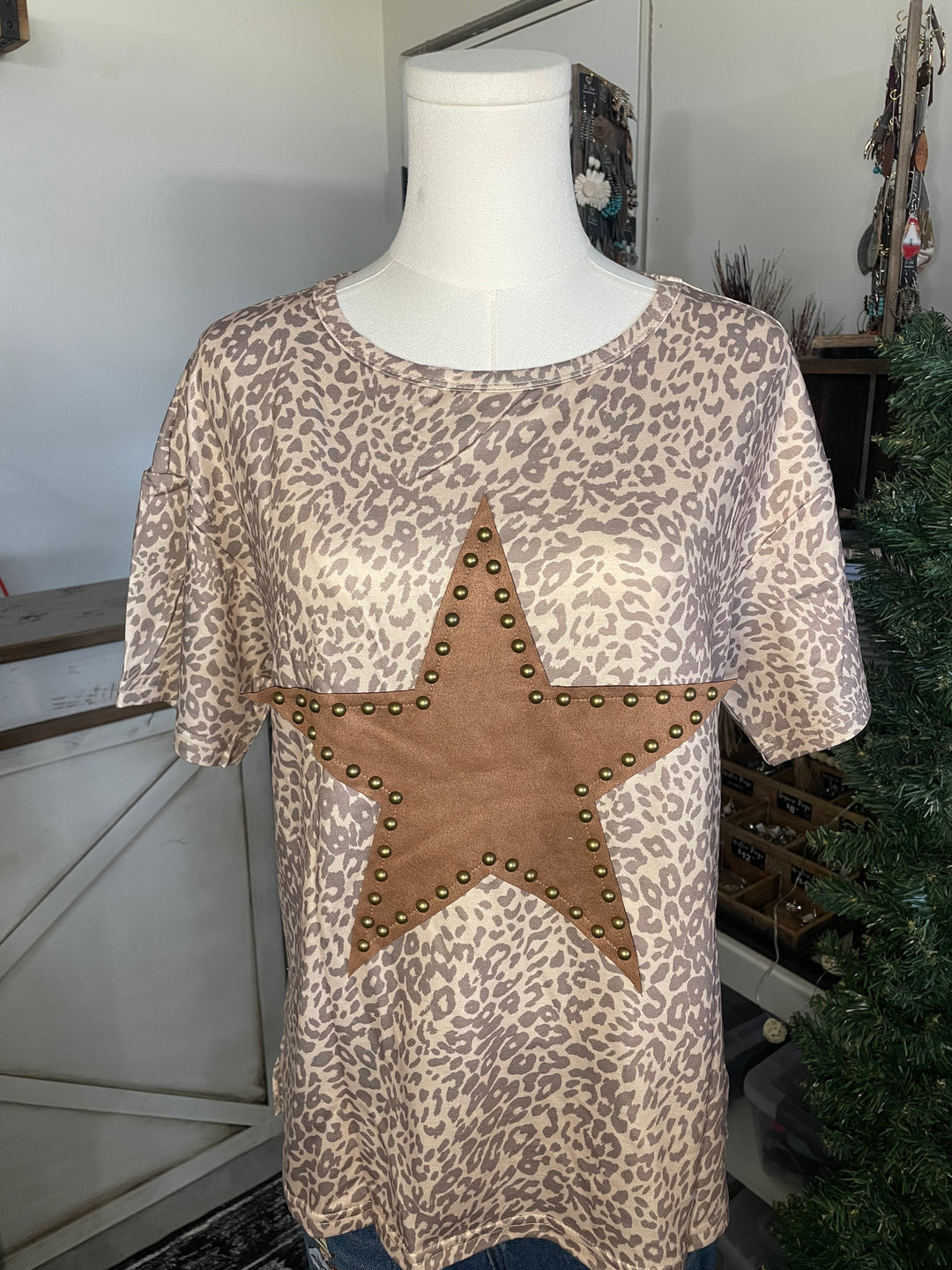 A short-sleeve crewneck top displayed on a white mannequin torso. The shirt features a beige and brown leopard print pattern with a large brown faux-suede star applique on the front, decorated with small metallic studs.
