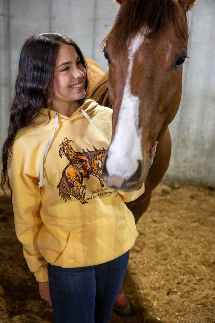 A woman wearing a yellow hoodie with a cowgirl riding a horse graphics, standing next to a real horse in a stable.