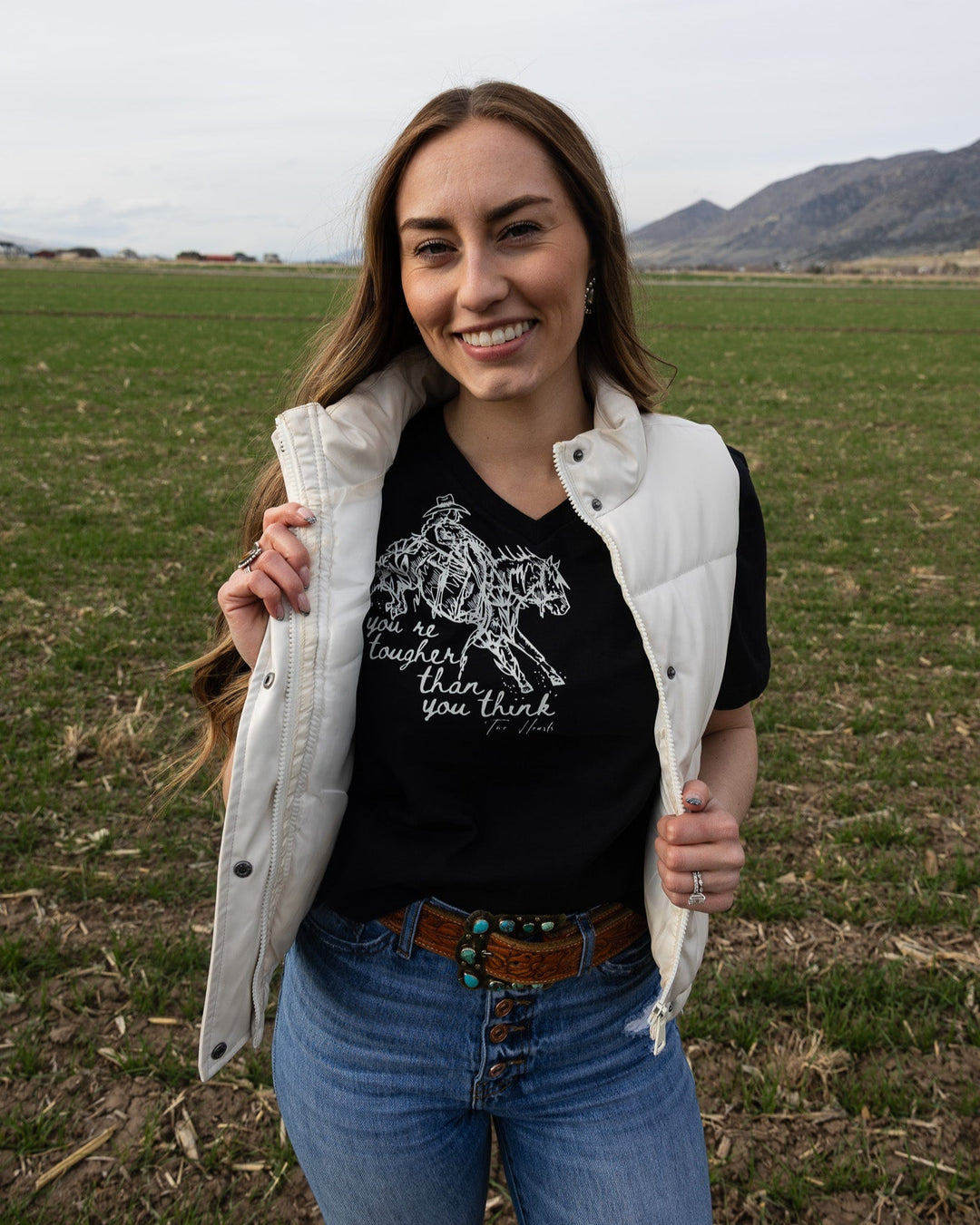 A person wearing a black V-neck tee with a graphic of a cowgirl riding a bucking horse and text, paired with blue jeans and a light-colored jacket.