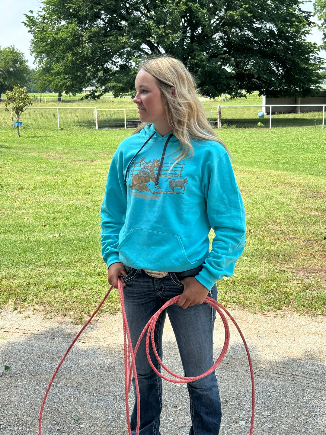 A person wearing a blue hoodie with a graphic print of a roper on a horse, standing in an outdoor setting with a lasso in hand.