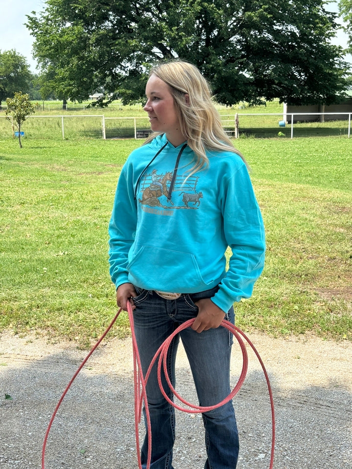 A person wearing a blue hoodie with a graphic print of a roper on a horse, standing in an outdoor setting with a lasso in hand.