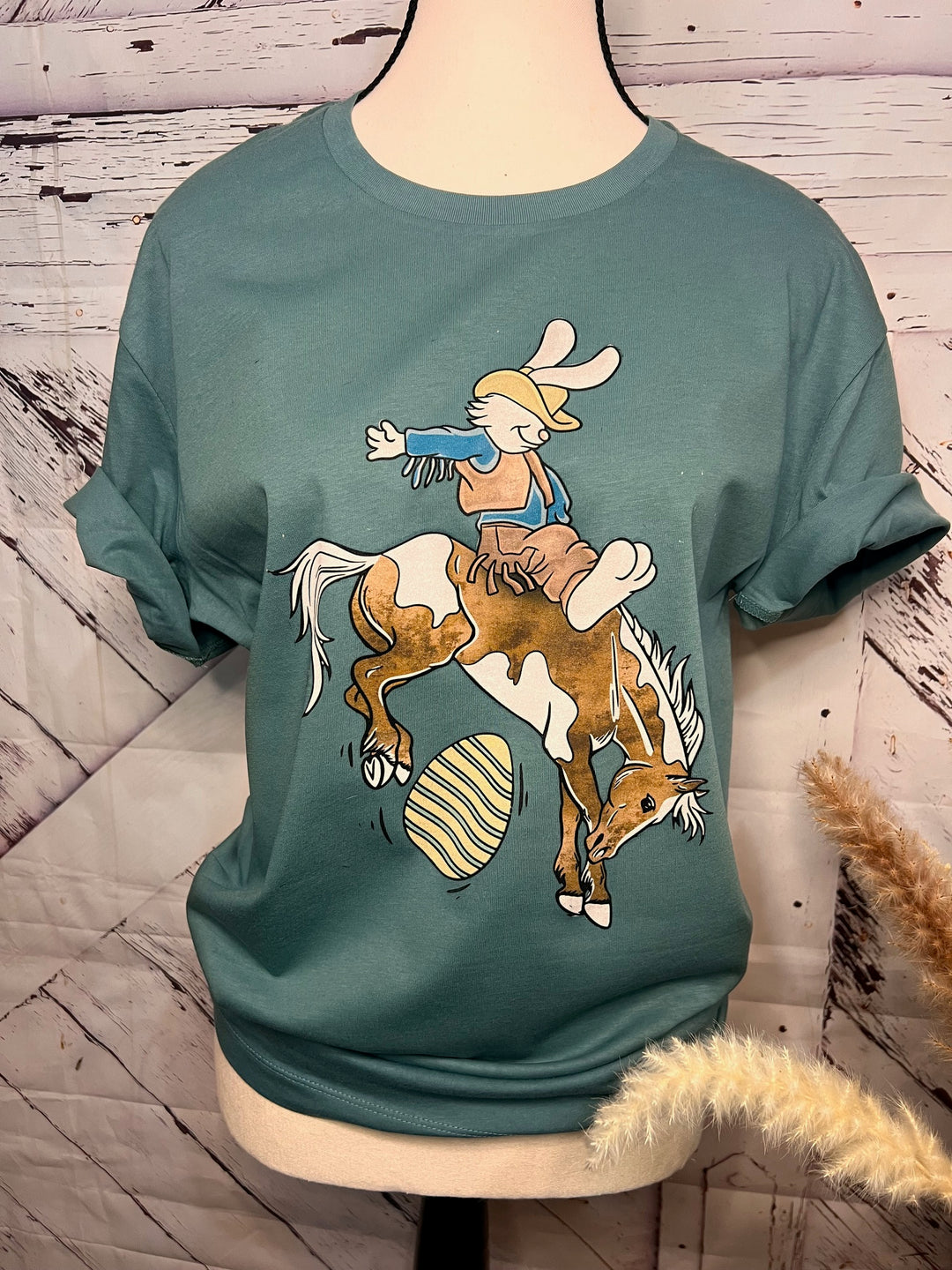 A graphic tee with a beige color featuring a graphic of a bucking bronco with an Easter bunny riding on top, displayed on a mannequin.