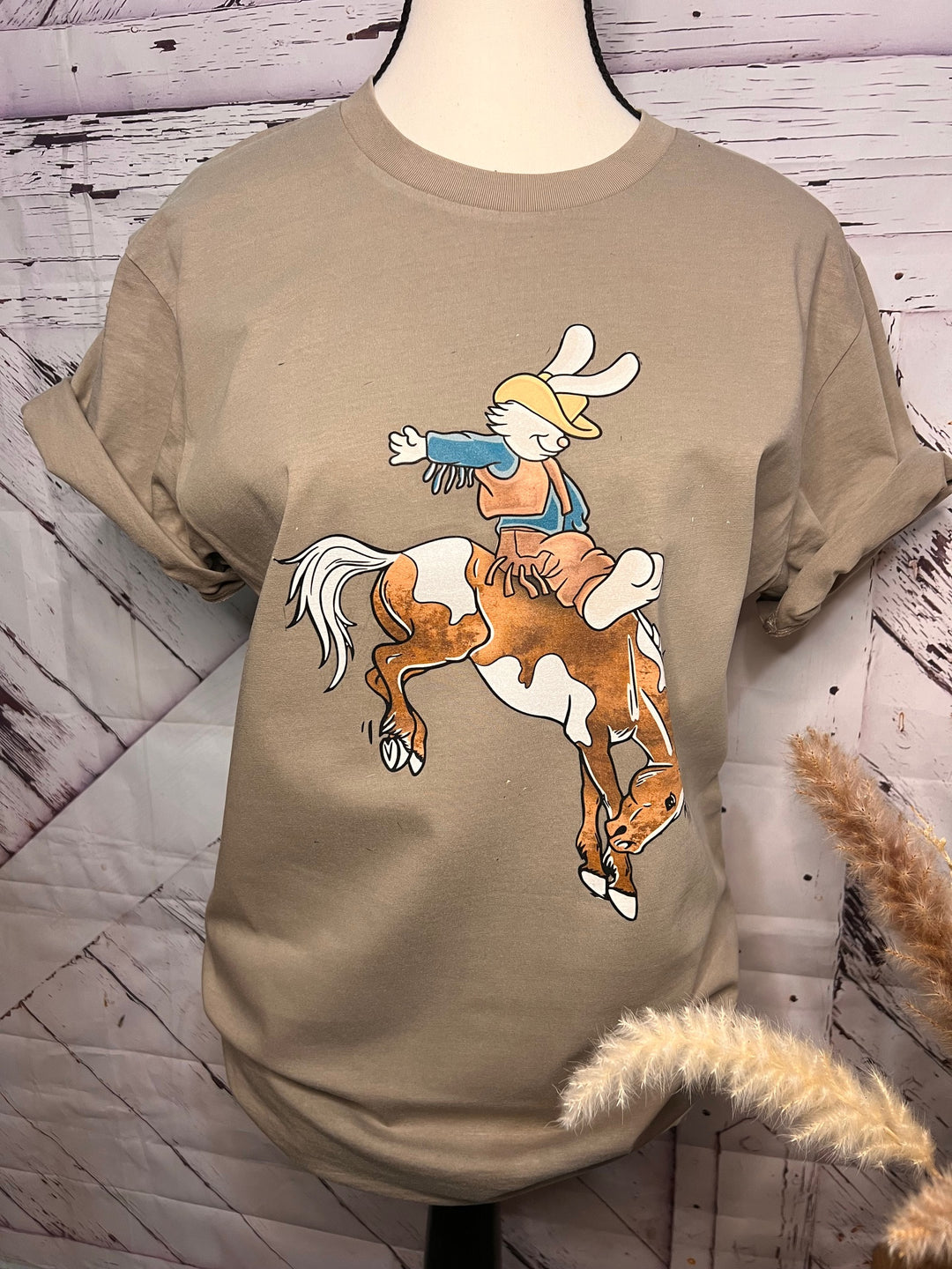 A graphic tee with a beige color featuring a graphic of a bucking bronco with an Easter bunny riding on top, displayed on a mannequin.