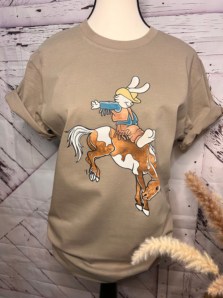 A graphic tee with a beige color featuring a graphic of a bucking bronco with an Easter bunny riding on top, displayed on a mannequin.
